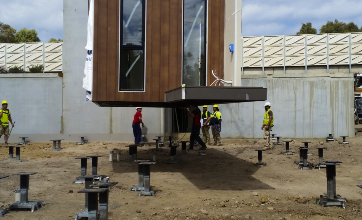 A graphic illustration of a home panel -- a wall with windows -- being dropped onto steel foundations as workers watch.